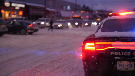 Coche-De-Policía-Con-Luces-Encendidas,-Bloqueando-La-Carretera-Mientras-Los-Coches-Conducen-A-Través-De-La-Nieve