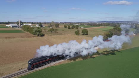 Vista-Aérea-De-Un-Tren-De-Vapor-Que-Sopla-Humo-Y-Vapor-Mientras-Viaja-Por-El-Campo-Seguido-Por-Delante-Del-Tren