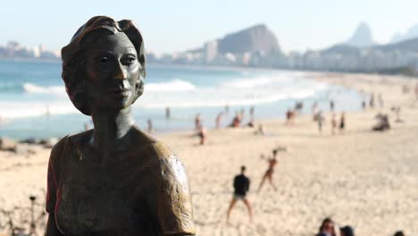 Estatua-De-Bronce-De-Clarice-Lispector,-Periodista-Y-Escritora-Ucraniana-Nacida-En-1920-Y-Naturalizada-Brasileña-Con-Gente-Haciendo-Actividades-De-Ocio-En-La-Playa-De-Copacabana-Al-Fondo