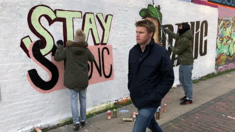 Graffiti-Künstler-Sprühen-Eine-Figur-Mit-Einer-Chirurgischen-Gesichtsmaske-Und-Der-Anti-Coronavirus-Botschaft-„Bleib-Stark-Und-Keine-Panik“-Auf-Eine-Wand,-Während-Menschen-Vorbeigehen