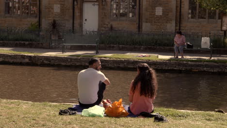 Durante-La-Pandemia-De-Covid-19,-Se-Ve-A-Una-Pareja-Con-Otras-Personas-Disfrutando-De-Un-Día-Soleado-Al-Lado-De-Un-Canal-En-Bourton-on-the-water,-Cotswold,-Reino-Unido,-Mientras-Mantienen-El-Distanciamiento-Social