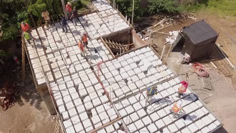 Vista-Aérea-De-Los-Trabajadores-De-La-Construcción-Que-Trabajan-En-El-Techo-Vertiendo-Cemento-Para-Construir-Una-Estructura-De-Techo-O-Piso-Usando-Chalecos-Naranjas