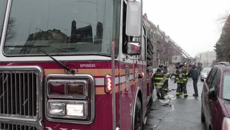 Camión-De-Bomberos-Con-Luces-Intermitentes-Estacionarias-En-La-Calle-Brooklyn-Bajo-Fuertes-Nevadas-Con-Un-Grupo-De-Bomberos---Plano-Medio