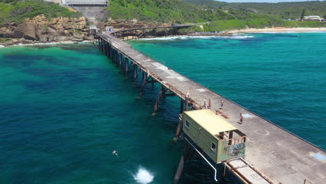 Young-male-jumping-off-pier-into-sea-with-friends-watching,-aerial-reveal