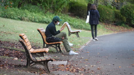 Tipo-Con-Mascarilla-Sentado-Y-Leyendo-Un-Libro-Mientras-Escucha-Música-En-Un-Banco-De-Madera-En-El-Parque-Durante-Una-Pandemia