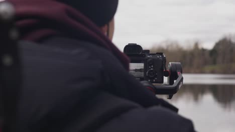 Man-on-dock-filming-the-landscape-with-his-sony-DSLR-camera-mounted-on-a-ziyun-gimbal-crane-stabilizer-at-lake-waterfront-with-blurry-clouds-trees-water-in-background