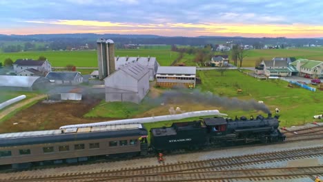 Una-Vista-Aérea-De-Un-Antiguo-Tren-De-Vapor-Restaurado-Y-Vagones-De-Pasajeros-Que-Se-Acercan-Con-Humo-Y-Vapor-Tirando-Hacia-El-Patio-Y-La-Estación-Vistos-Por-Un-Dron