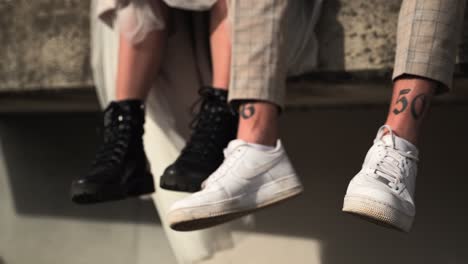 Close-up-of-wedding-couple-in-black-boots-and-white-nike-sneaker-sitting-on-the-edge-of-stone-wall-warehouse-gate-urban-environment