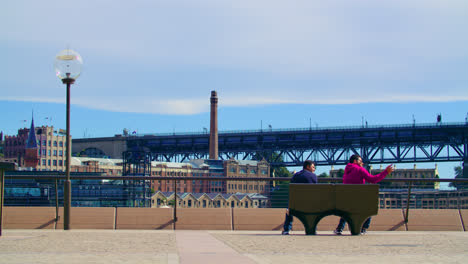 Two-Men-Sitting-At-The-Bench-With-The-Rocks-Buildings-In-The-Background-In-Sydney,-NSW,-Australia