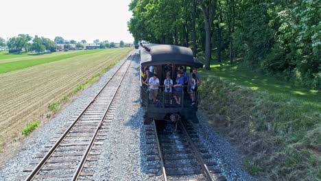 Vista-Aérea-De-Un-Dron-Que-Viene-A-La-Parte-Trasera-De-Un-Antiguo-Vagón-De-Pasajeros-De-Tren-Con-Pasajeros-En-La-Parte-De-Atrás-Saludando-Al-Dron