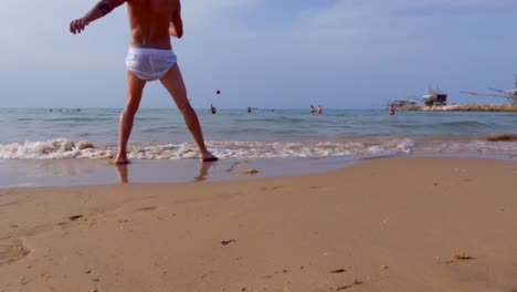 Punto-De-Vista-De-La-Superficie-Del-Suelo-De-ángulo-Bajo-Del-Niño-Pateando-La-Pelota-Roja-Desde-La-Orilla-Del-Mar-Hasta-El-Agua-De-Mar-Con-El-Hombre-Saltando-Para-Salvar