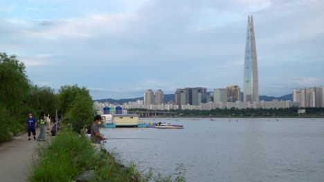 Hombre-Pescando-En-La-Orilla-Del-Río-Han-Con-El-Paisaje-Urbano-De-Seúl-Y-La-Torre-Mundial-Lotte-En-Corea-Del-Sur