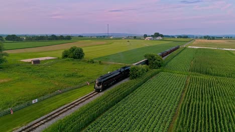 Una-Vista-Aérea-De-Un-Tren-De-Vapor-Que-Se-Acerca-Volando-Viajando-A-Través-De-Tierras-De-Cultivo-Y-Campos-De-Maíz-Que-Soplan-Humo-En-Un-Día-Soleado-De-Verano