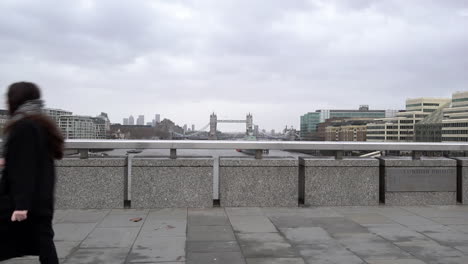 La-Gente-Cruza-El-Puente-De-Londres-Y-Pasa-El-Puente-De-La-Torre-Y-El-Río-Támesis-En-El-Fondo-En-Un-Día-Frío,-Gris-Y-Ventoso-En-Cámara-Lenta