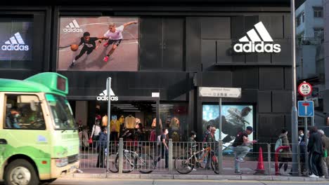 Los-Peatones-Pasan-Frente-A-La-Marca-Multinacional-Alemana-De-Ropa-Deportiva-Adidas,-La-Tienda-Oficial-Y-El-Logotipo-En-Hong-Kong