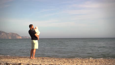 Padre-Sosteniendo-Amorosamente-A-Un-Bebé-Pequeño-Mientras-Mira-El-Océano-En-La-Playa