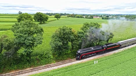 Una-Vista-Aérea-De-Las-Tierras-Agrícolas-Amish-Con-Una-Sola-Vía-Férrea-Y-Un-Tren-De-Pasajeros-A-Vapor-Acercándose