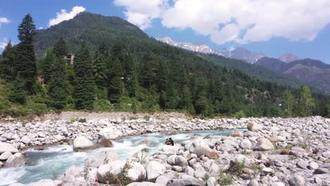 Aerial-shot-of-water-and-pine-tree-forest-Aerial-shot-of-water-stream-flowing-near-the-rocks-with-a-mountain-in-the-background