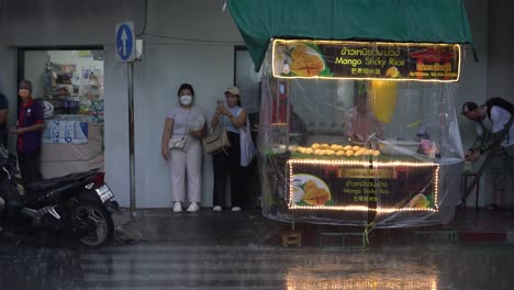 Gente-Esperando-Que-La-Lluvia-Se-Detenga-Junto-Al-Puesto-De-Arroz-Pegajoso-De-Mango