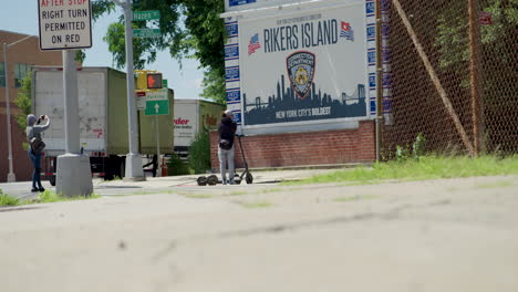 Personas-En-Scooters-Eléctricos-Toman-Una-Foto-Frente-Al-Cartel-De-La-Cárcel-De-Rikers-Island-En-Nyc