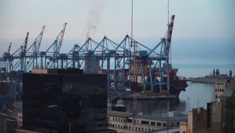 Edificios-De-Valparaíso-En-El-Frente,-Buque-De-Carga-De-Contenedores-Atracado-Esperando-Ser-Cargado-Cerca-De-Grúas-En-El-Puerto-Marítimo,-Chile