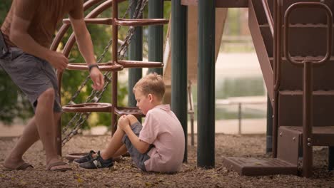 Weinender-Verletzter-Junge-Auf-Dem-Spielplatz-Im-Park-Mit-Knieverletzung,-Vater-Kommt,-Um-Ihm-Ein-Pflaster-Zu-Geben-Und-Ihm-An-Einem-Hellen-Sommertag-Aufzuhelfen