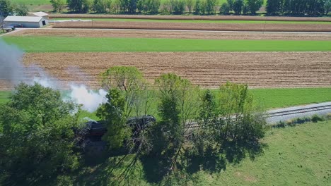 Una-Vista-Lateral-De-Un-Dron-De-Un-Tren-De-Pasajeros-De-Vapor-Antiguo-En-Cámara-Lenta-Que-Sopla-Humo-Y-Vapor-Que-Viaja-A-Través-De-Campos-De-Maíz-Fértiles-Y-Viaja-A-Través-De-árboles-En-Un-Día-Soleado-De-Verano