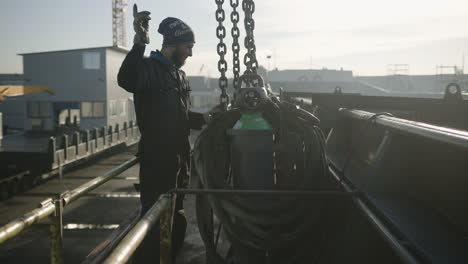 Mann,-Der-Einen-Kranhaken-An-Einem-Gastank-Befestigt-Und-Mit-Seinen-Fingern-Signalisiert,-Ihn-An-Einem-Frachtschiff-Im-Hafen-Anzuheben