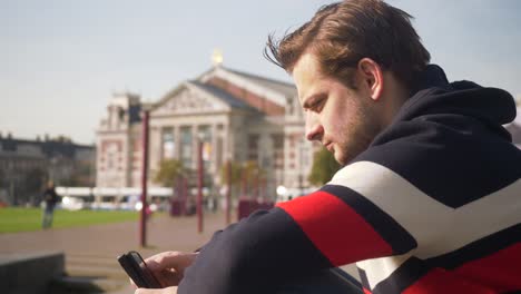 Hübscher-Junger-Mann,-Der-An-Einem-Sonnigen-Tag-Auf-Dem-Handy-In-Der-Nähe-Der-Konzerthalle-In-Amsterdam-Tippt