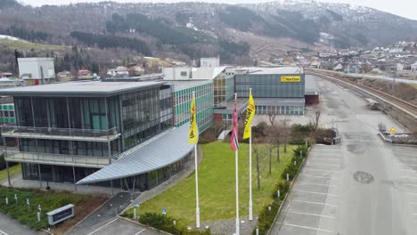 Nordische-Fabrikanlagen-Und-Flaggen-In-Moi-Norwegen---Aufsteigen-Aus-Der-Luft-Vor-Wehenden-Flaggen,-Bevor-Der-Panoramablick-Auf-Die-Fabrik-Freigelegt-Wird---Fenster--Und-Türenfabrik