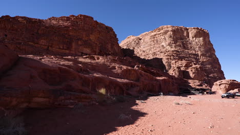 Massive-Red-Rock-Mountains-in-the-Middle-of-Wadi-Rum-Desert-on-a-Sunny-Day-With-Tourists-Climbing-on-the-Hill-and-Bedouins-Jeps-Waiting