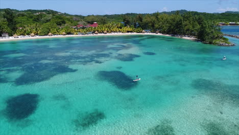 Vista-Aérea-En-Cámara-Lenta-De-Personas-Haciendo-Paddleboarding-Cerca-De-La-Playa-De-Un-Resort-En-El-Mar-Caribe-Hondureño