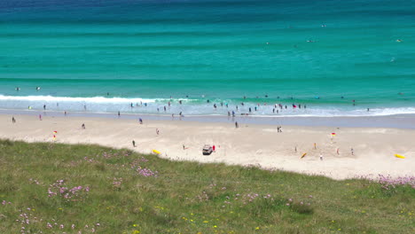 Ocean-waves-along-the-wide-sandy-shore-at-Sennen-Cove-in-Cornwall-with-surfers-and-tourists-walking-on-the-beach