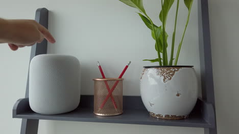 Toma-Estática-De-Un-Hombre-Tocando-Dos-Veces-Un-Homepod-De-Apple-En-La-Parte-Superior-De-Una-Estantería-De-Aspecto-Moderno-Junto-A-Un-Portalápices---Planta