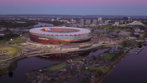 Vista-Aérea-Del-Estadio-Optus-De-Perth-A-La-Hora-Azul-Con-Colores-Degradados-En-El-Cielo-Y-Gente-Caminando-Hacia-El-Partido,-Mientras-La-Cámara-Se-Mueve-Hacia-La-Derecha-Y-Alrededor-Del-Estadio