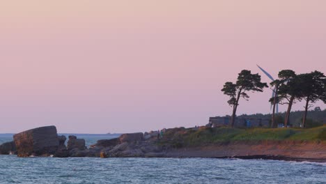 Distant-people-enjoying-romantic-sunset-by-the-sea-with-a-wind-turbine-in-the-background,-wide-shot