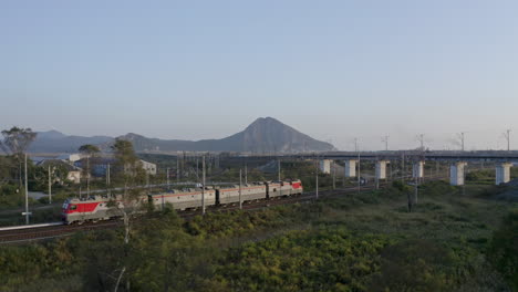 Russian-railway-train-locomotives-move-through-the-fields-with-mountains-in-the-background,-approaching-a-car-bridge-over-the-railway,-on-the-sunset,-Russian-Federation