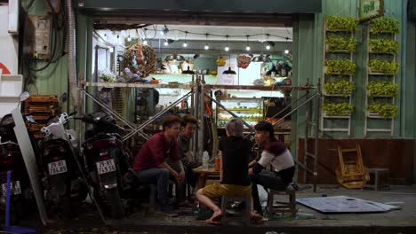 Four-young-men-have-some-drinks-at-a-sidewalk-table-on-a-small-restaurant-bar-at-night,-mopeds-are-parked-to-their-left,-Locked-shot