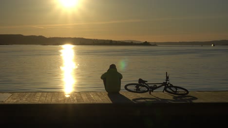 Silueta-Masculina-Sentada-Al-Lado-De-La-Bicicleta-En-El-Puerto,-Viendo-La-Puesta-De-Sol-Sobre-El-Fiordo-De-Oslo