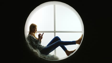 Mädchen-Sitzt-Im-Runden-Fenster-Mit-Smartphone