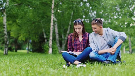 Young-Man-And-Woman-Sitting-On-Grass-Looking-at-Book