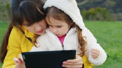Young-Mother-With-A-Funny-Little-Girl-In-A-White-Sweatshirt-With-A-Hood-Playing-On-A-Tablet-In-The-S