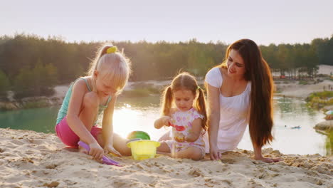 Junge-Mutter-Spielt-Im-Sand-Mit-Zwei-Töchtern-Bei-Sonnenuntergang-Auf-Dem-Hintergrund-Des-Schönen-Sees