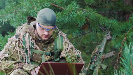 Portrait-Of-Military-Man-Sitting-In-The-Woods-Dressed-In-Camouflage-It-Uses-Laptop