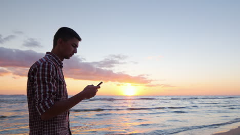 Young-Lonely-Man-Walking-Along-Seashore-At-Sunset-Typing-On-Phone
