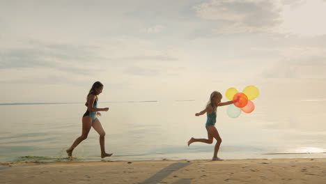 Zwei-Schwestern,-Die-Zusammen-Am-Strand-Spielen,-Ein-Mädchen-Läuft-Nach-Dem-Anderen-Und-Hält-Ballons-In-Zeitlupe
