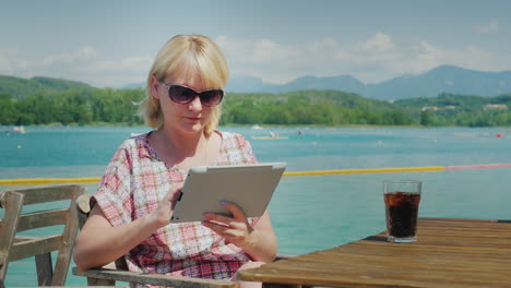 Tecnología-En-Vacaciones-Mujer-Con-Tableta-Sentado-En-Una-Mesa-En-Un-Café-En-El-Fondo-Del-Lago