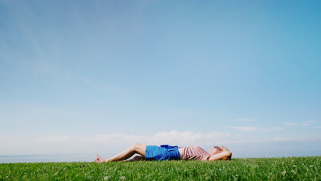 A-Young-Woman-Enjoys-Warmth-And-Summer-Lying-On-The-Green-Grass-Against-The-Blue-Sky