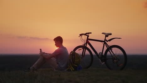 The-Teenager-Uses-A-Smartphone-Sits-Near-His-Bike-And-Backpack-At-Sunset