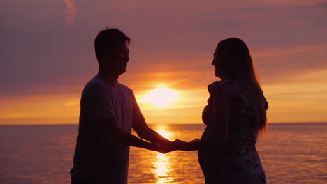 Seitenansicht-Eines-Asiatischen-Mannes,-Der-Die-Hände-Seiner-Schwangeren-Frau-Im-Hintergrund-Des-Sonnenuntergangs-über-Dem-Meer-Hält